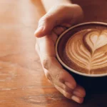 Zwei Hände umgreifen eine Tasse mit Kaffee. Darauf schwimmt ein Milchschaum Herz